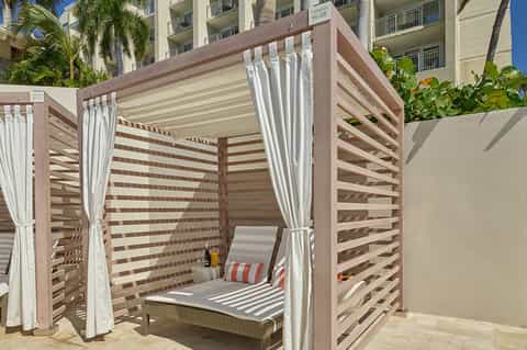 Outdoor cabana with brown and white striped pergola, white curtains, and lounge seating