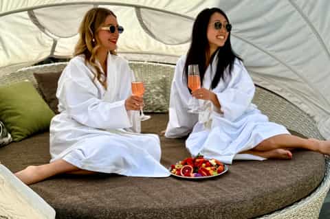 Two women in white robes holding champagne glasses in glamorous tent cabana with fruit platter