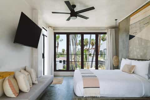 Modern resort bedroom with bed, seating area, flat-screen TV, and sliding glass doors opening to palm tree views