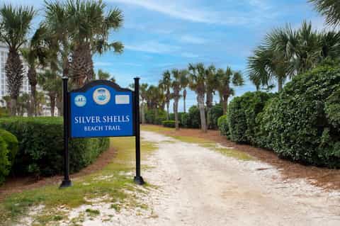 Sandy beach trail entrance marked by blue Silver Shells Beach Trail sign lined with palm trees