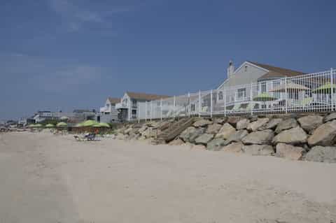 Beachfront homes with white railings on sandy shore protected by large rock breakwater
