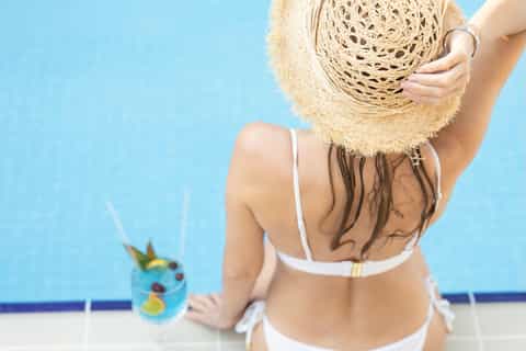 Woman in white swimsuit holding straw hat by pool with tropical drink on table