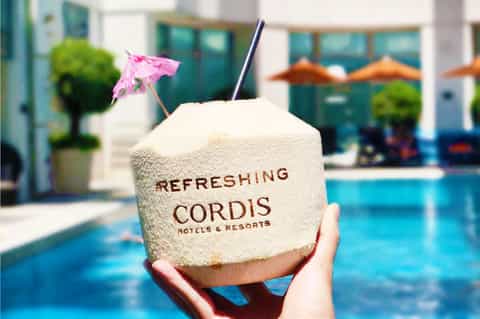 Coconut drink with pink umbrella and straw by resort pool with lounge chairs and umbrellas