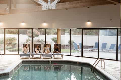 Indoor pool area with lounge chairs, large windows overlooking landscape, and exposed wooden beams