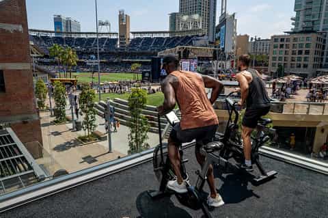 Two people on stationary bikes overlooking a baseball stadium in an urban setting