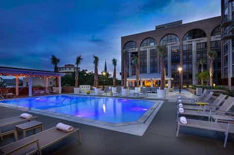 Illuminated resort pool at twilight with blue lighting, lounge chairs, palm trees, and brick building façade