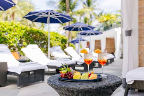Upscale poolside table with cocktails and fruit platter under blue umbrellas