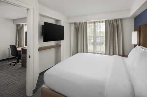 Hotel guest room with white bedding, wall-mounted TV, work desk, chair, and large windows with neutral curtains
