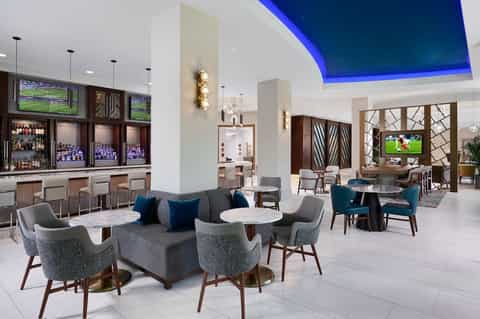 Modern hotel lobby lounge with blue-lit ceiling, contemporary furniture, bar, and flat-screen TVs