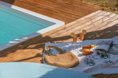 Poolside lounging area with turquoise pool, woven beach bag, cocktails, and magazines on towel
