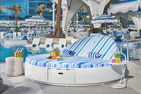Luxurious poolside cabana bed with blue striped cushions, umbrellas, and resort facilities in background