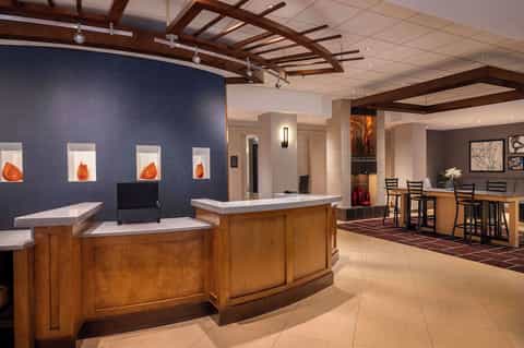 Hotel reception area with curved wooden desk, illuminated amber display wall, coffered ceiling, and bar lounge seating
