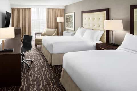 Hotel bedroom with two queen beds, beige headboards, dark wood furniture, and large windows