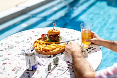 Hand holding beverage poolside while enjoying burger and fries overlooking swimming pool