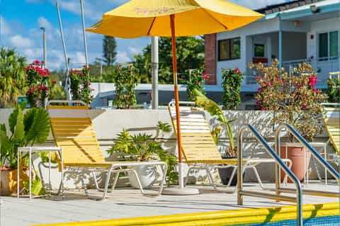 Bright poolside deck with yellow umbrella, lounge chairs, and flowering planters near building entrance