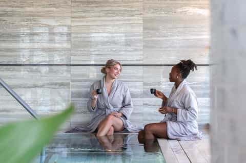 Two women in spa robes enjoying drinks poolside near marble wall and water feature