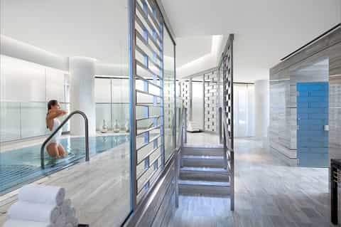 Modern indoor swimming pool with natural lighting, columns, person at pool edge, spa-like design aesthetic