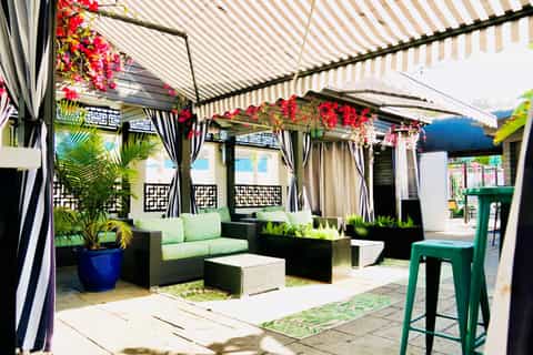 Covered outdoor patio with striped awning, hanging flowers, black furniture, and tropical plants
