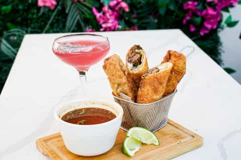 Cocktail and appetizers on white table with pink flowers, featuring fried shrimp and coral-colored drink