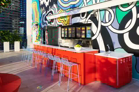 Colorful outdoor bar with vibrant street art mural, red counter seating, and beverage coolers