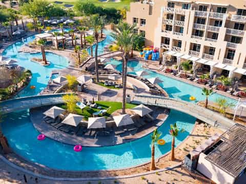 Aerial view of resort complex with curved swimming pools, palm trees, umbrellas, and multi-story buildings