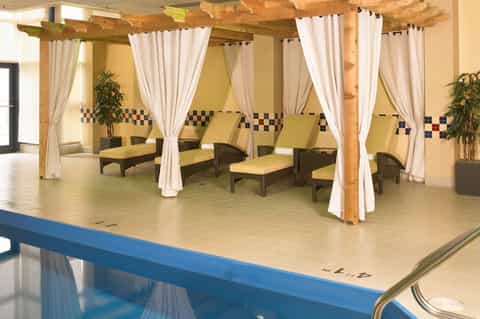 Spa relaxation area with wooden pergola, white curtains, yellow cushioned lounge chairs, and decorative tile border