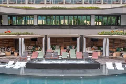 Resort pool deck with multi-colored loungers, cabanas with curtains, hot tub, and modern building