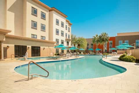 Resort pool area with turquoise water, white umbrellas, palm trees, and beige building facade