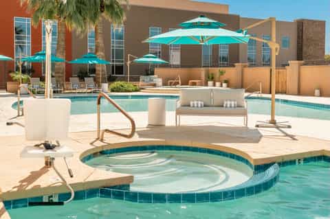 Resort pool area with turquoise umbrellas, lounge chairs, spa tub, and modern building