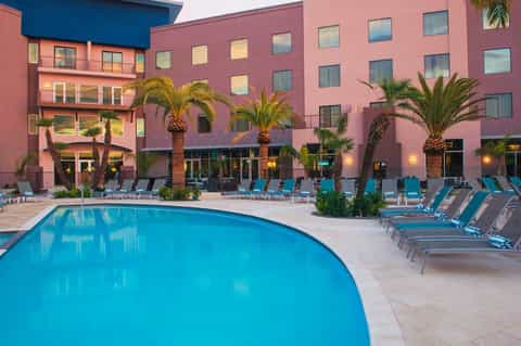 Resort pool area at dusk with pink building facade, palm trees, and lounge chairs
