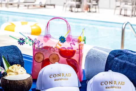 Colorful tropical drink in pink tote bag by resort pool with Conrad Nashville branded caps and towels