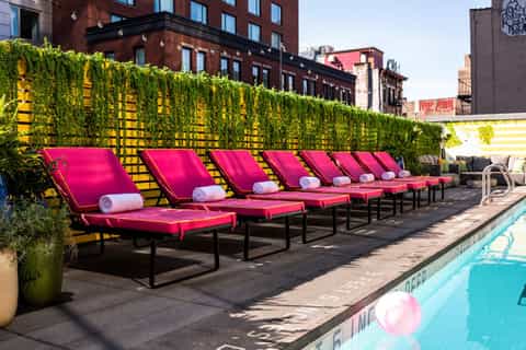 Urban rooftop pool deck with hot pink loungers, ivy wall, city buildings, and turquoise pool