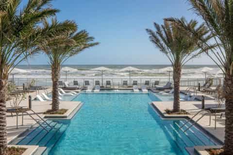 Beachfront resort pool with palm trees, white lounge chairs, ocean waves, and clear sky