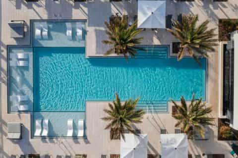 Aerial view of turquoise rectangular pool surrounded by white deck, palm trees, and modern buildings