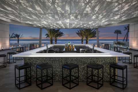 Rooftop bar with panoramic ocean and sunset views, perforated ceiling, modern counter seating, and palm trees