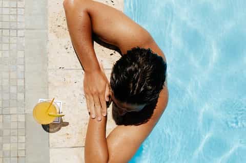 Shirtless man in pool holding tropical drink, overhead view from pool deck