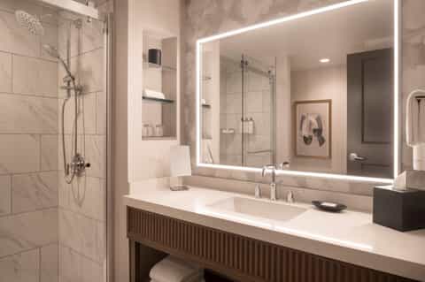 Modern bathroom with marble tiles, illuminated mirror, glass shower enclosure, and dual sinks