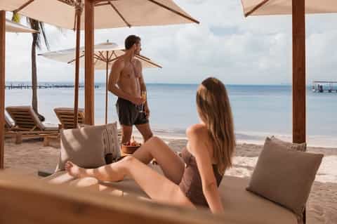Beachfront lounge with couple relaxing under umbrellas on white daybeds overlooking calm turquoise water and docks
