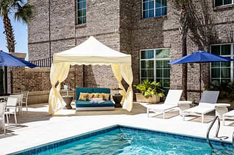 Poolside cabana with cream curtains and teal cushions beside brick building with blue umbrellas