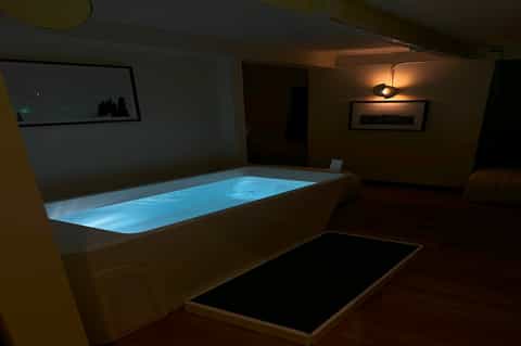 Indoor spa area with illuminated rectangular hot tub and black mat on wooden floor with accent lighting