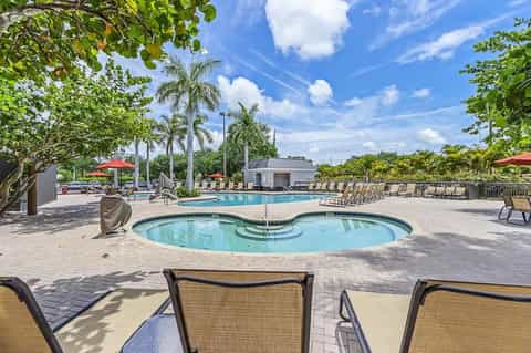Resort pool area with spa, palm trees, red umbrellas, and lounge seating on sunny day
