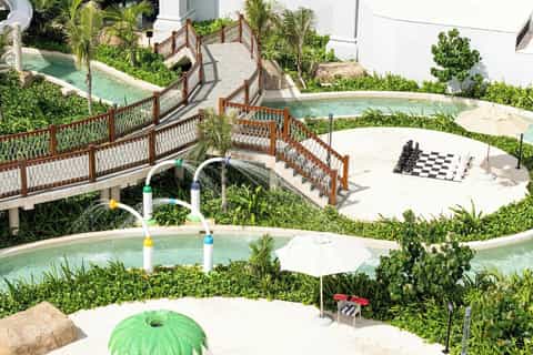 Multi-level resort grounds with pools, wooden bridges, palm trees, and shade umbrellas