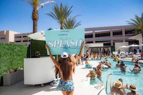 Woman holding turquoise splash sign at busy pool party with crowded swimming pool and palm trees