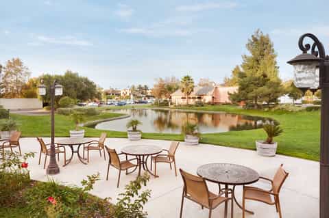 Outdoor patio with round tables, chairs, and decorative lamps overlooking pond and residential area