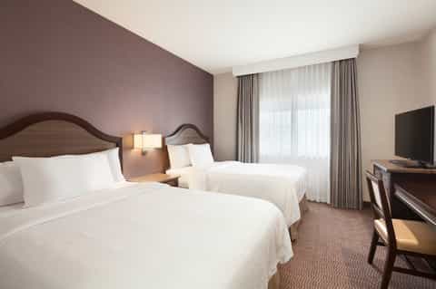 Traditional hotel room with two queen beds, mauve accent wall, and warm wood furnishings