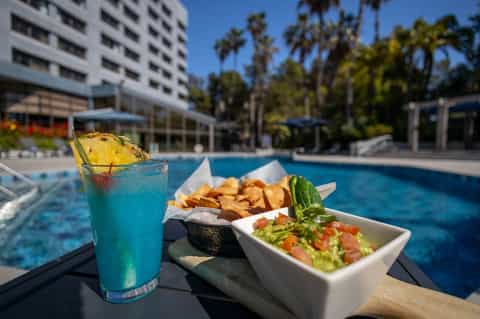 Colorful tropical cocktail and food plate poolside with resort building and palm trees in background