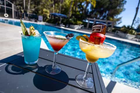 Three colorful cocktails displayed poolside with blue water and lounge chairs visible in background
