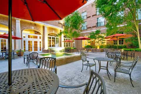 Illuminated courtyard with brick architecture, fountain, red umbrellas, and metal chairs at dusk