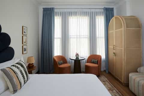 Modern hotel bedroom with blue curtains, brown armchairs, white bedding, and natural light