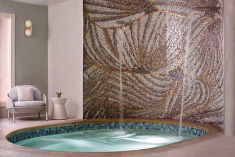 Luxury spa indoor hot tub surrounded by mosaic tile work, white wicker chair, and decorative wall sconce